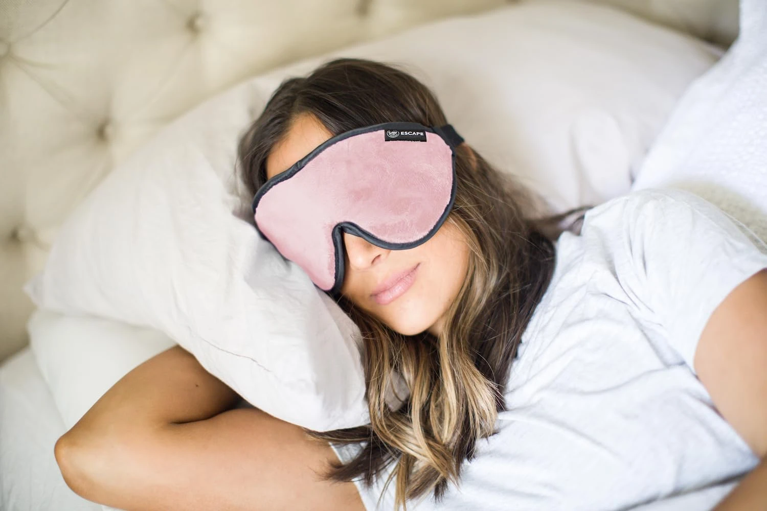 Dream Essentials Escape Luxe sleep mask in pink worn by a traveler in bed, ESCAPE logo tag visible